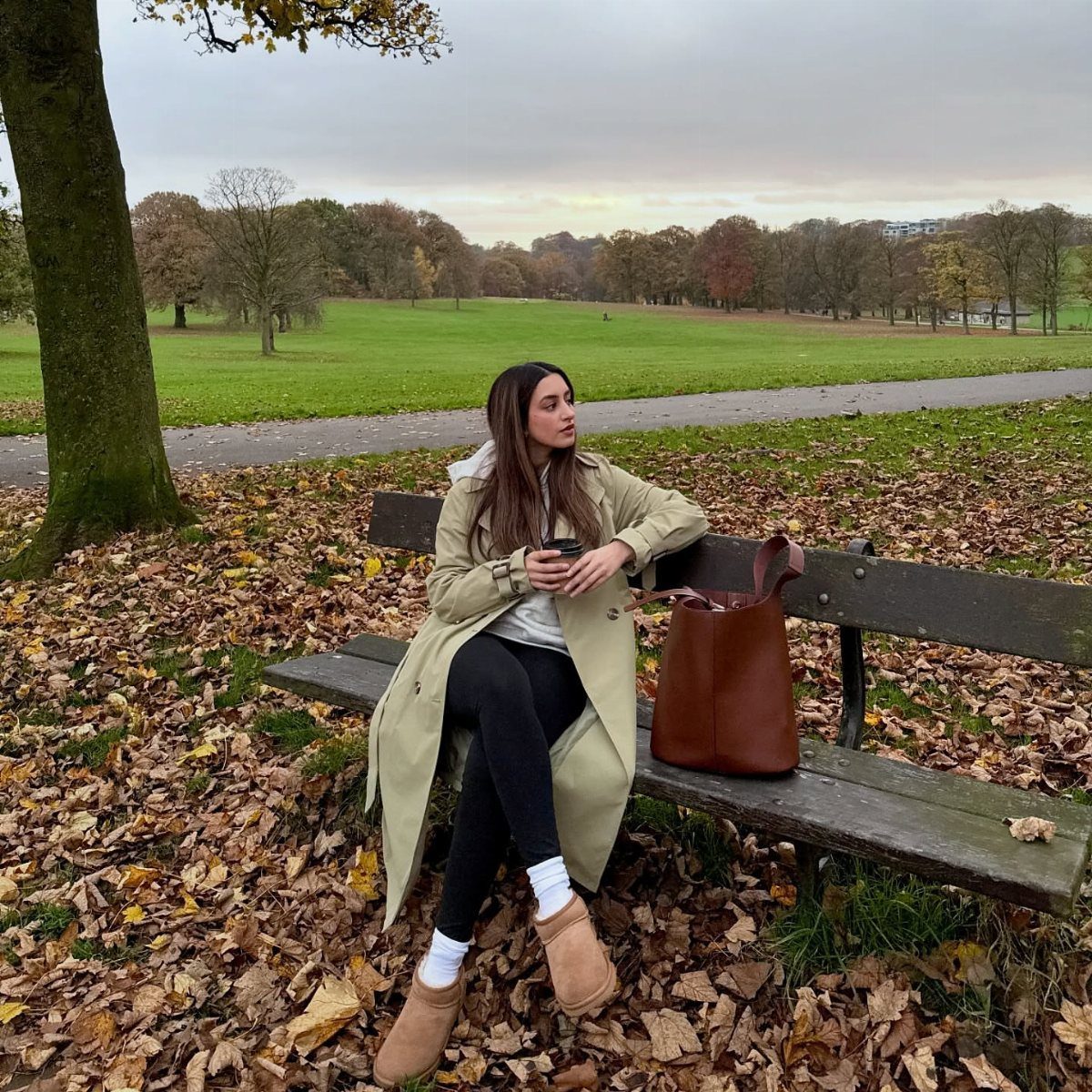 Woman in a beige trench coat and black leggings sitting on a park bench.