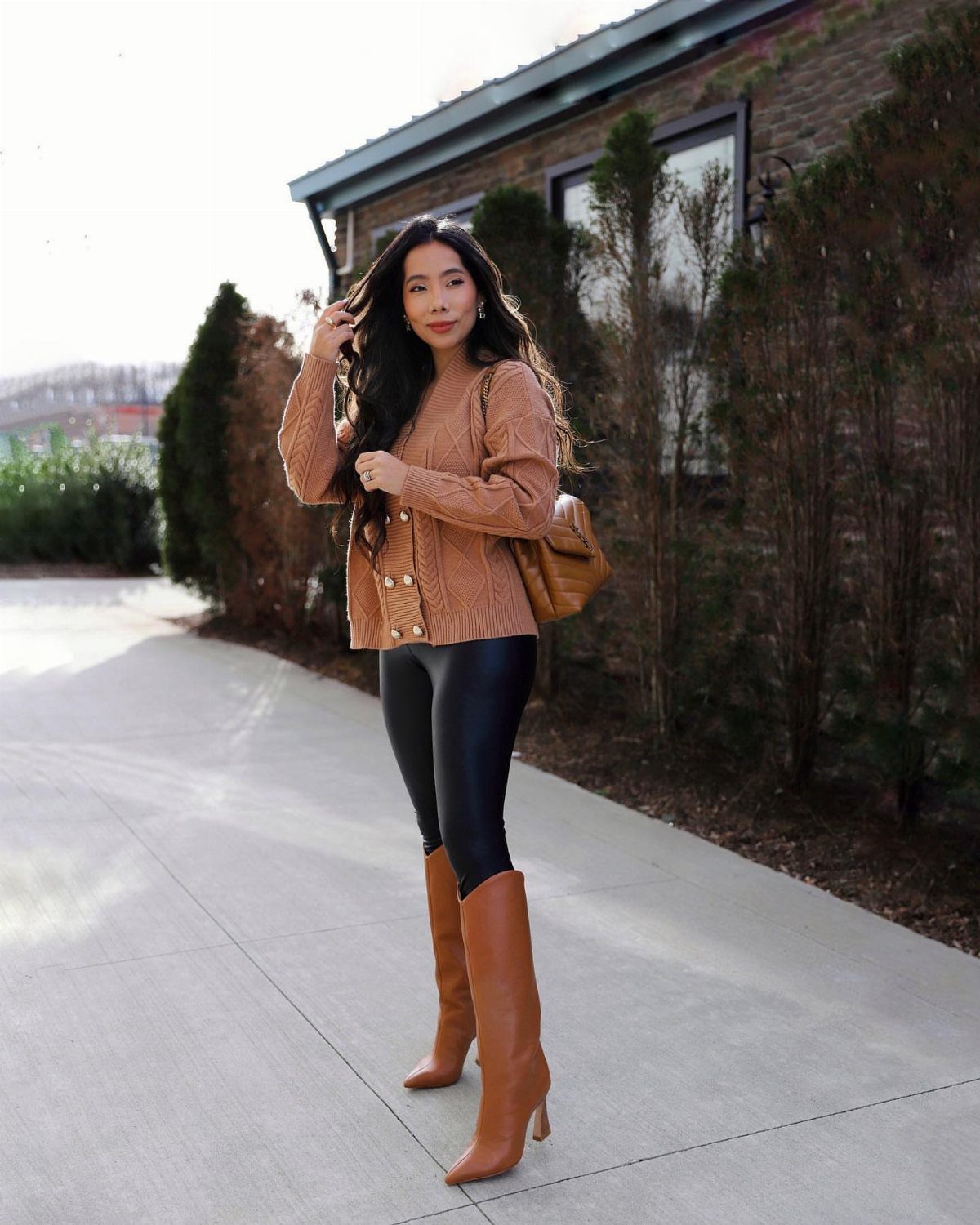 A woman wearing a tan knit sweater, black leather leggings, and tall tan boots outdoors.
