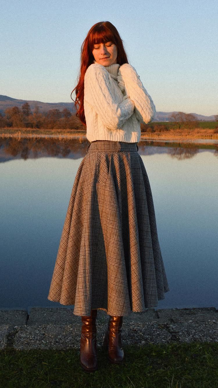 A woman wearing a white knit sweater and a long patterned skirt standing by a lake.
