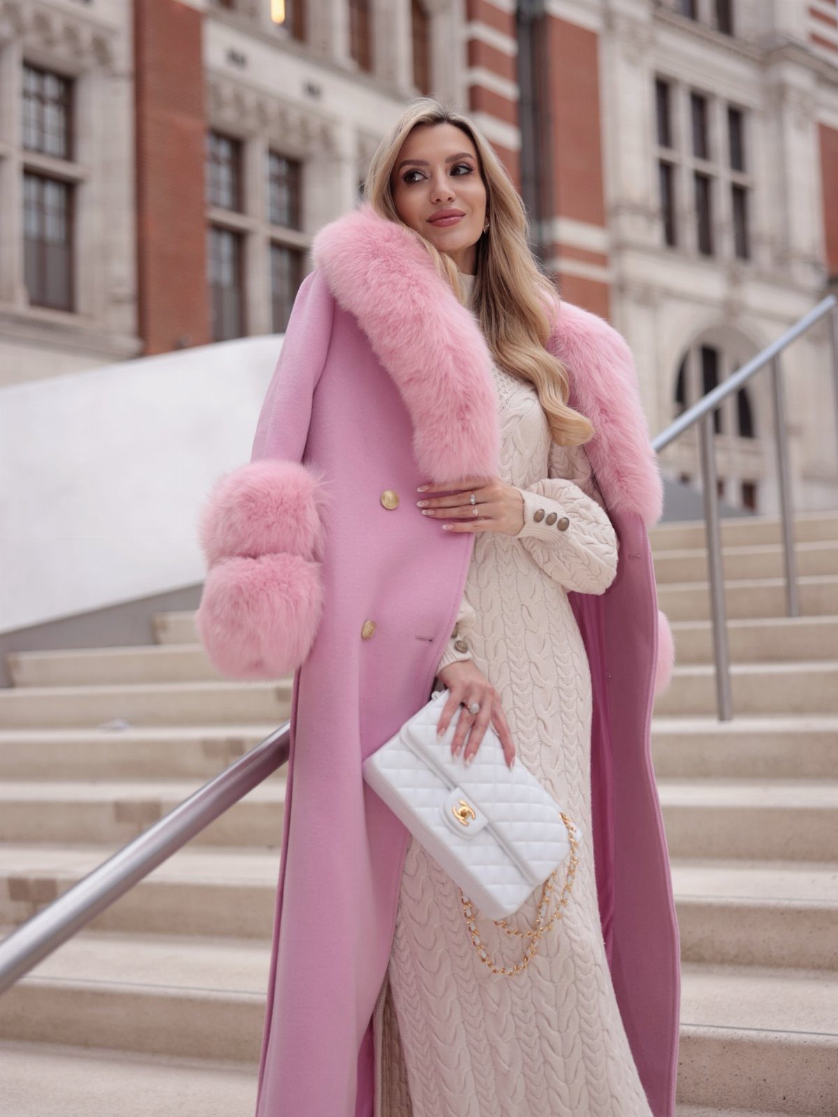 A woman wearing a pastel pink fur-trimmed coat and a cream knit dress standing on stairs.