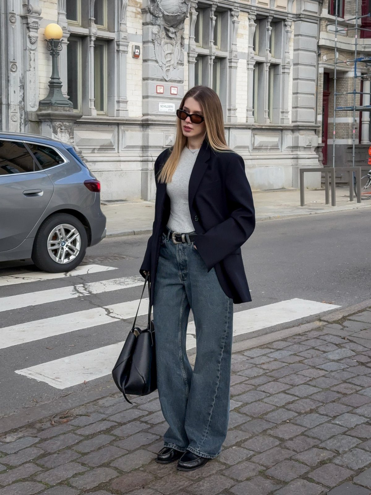 A woman in a black blazer, white top, and wide-leg jeans standing on a city street.