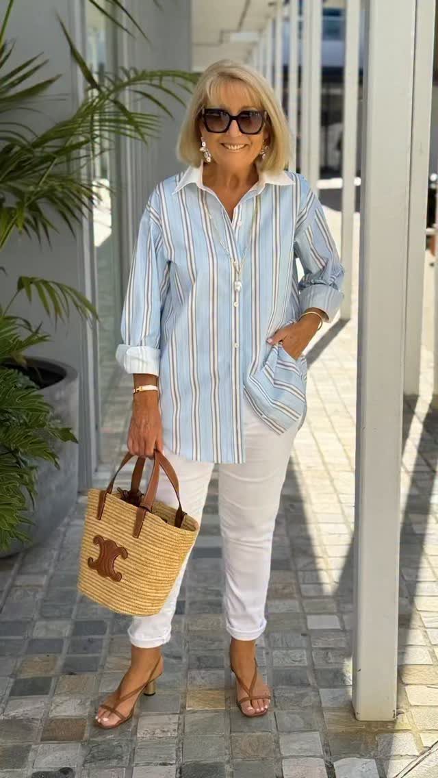 A blonde woman in a striped shirt and white trousers holding a straw bag.
