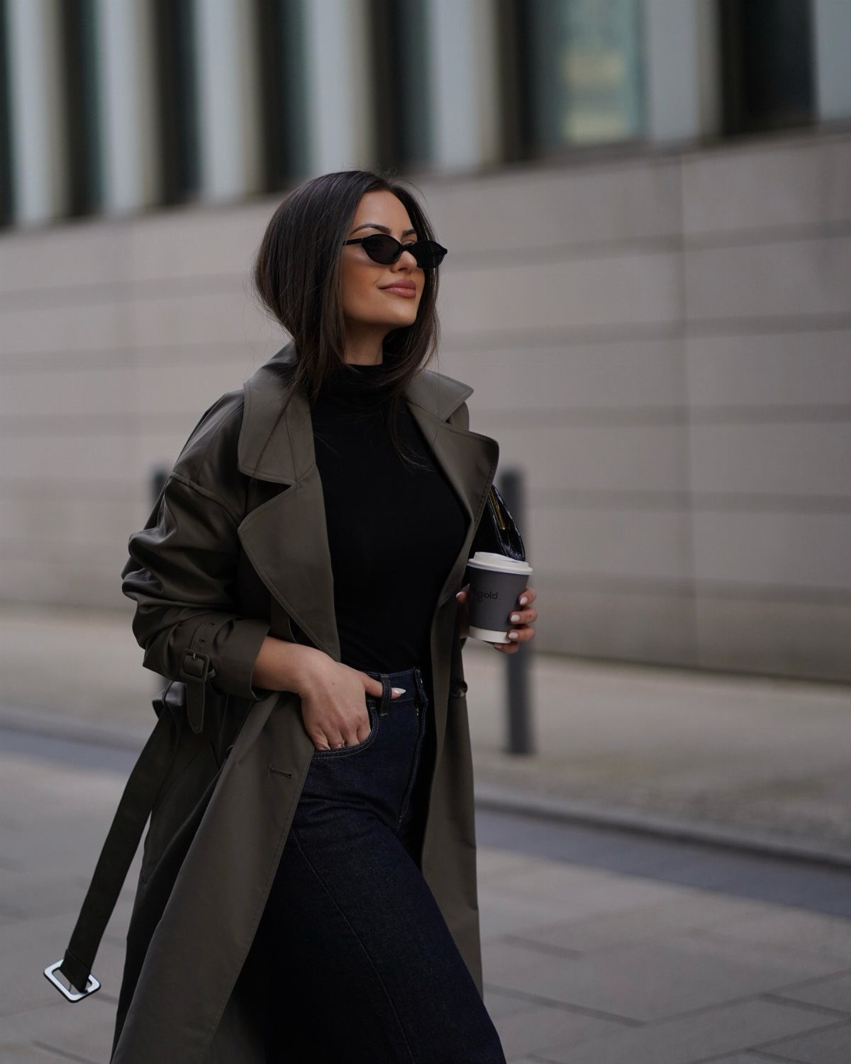Woman in an olive green trench coat and black turtleneck walking with a coffee cup.
