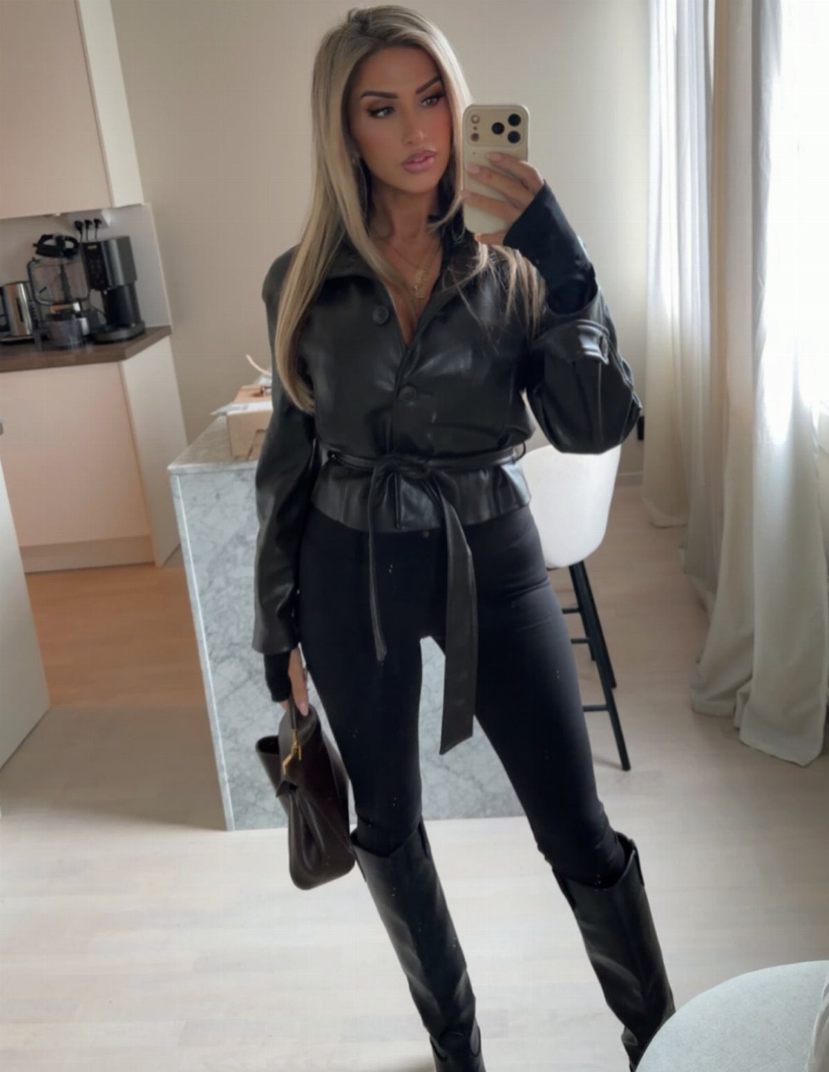 Blonde woman in a black leather jacket, black tights, and tall black boots posing for a selfie.