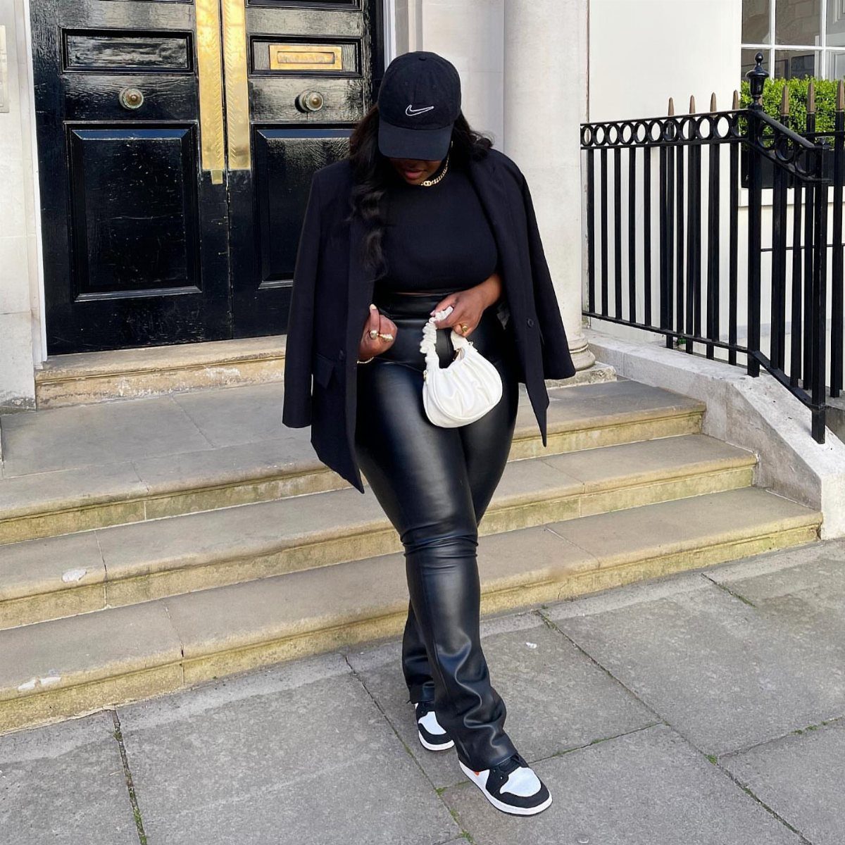 Person wearing black leather pants, black blazer, and black and white sneakers standing on stone steps.