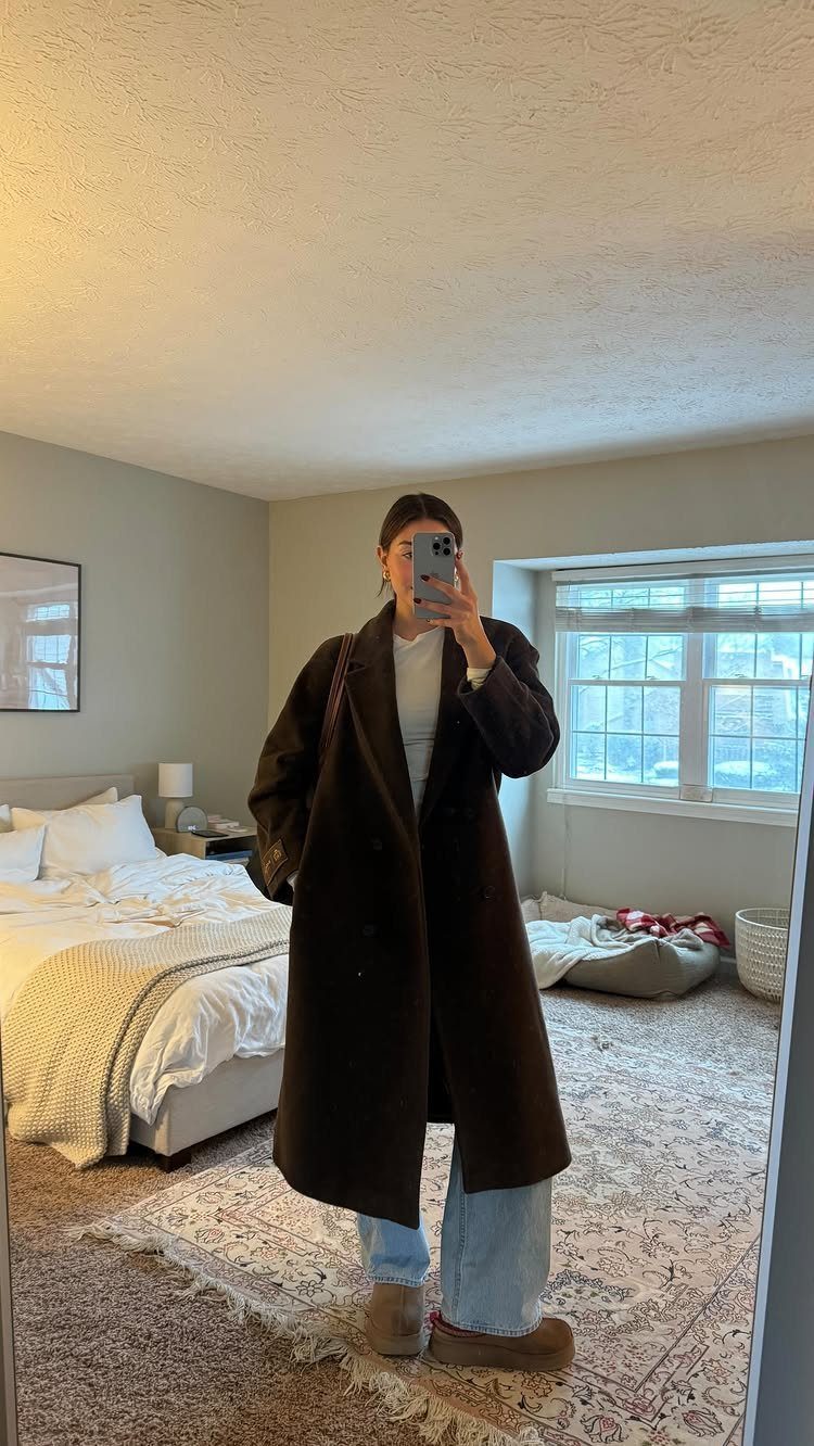 A person in a long brown wool coat and light blue jeans taking a mirror selfie.