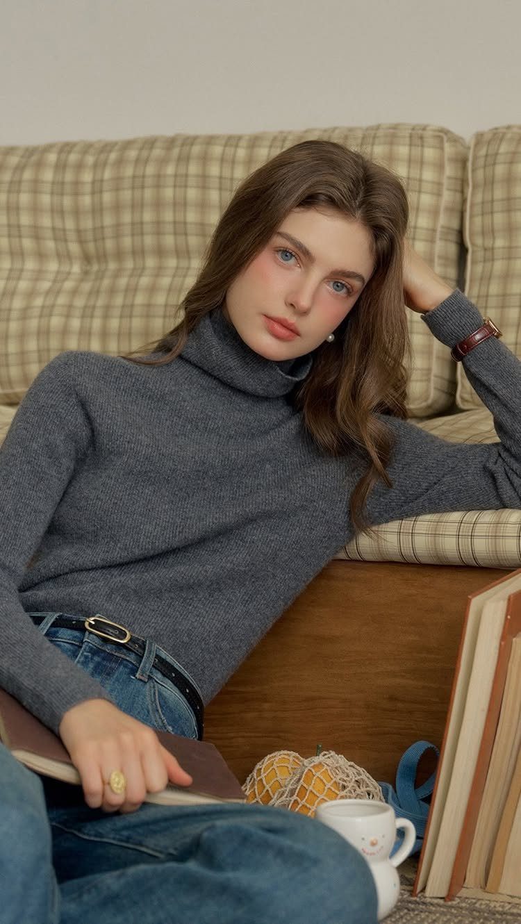 A woman wearing a grey turtleneck sweater and blue jeans sitting on the floor.