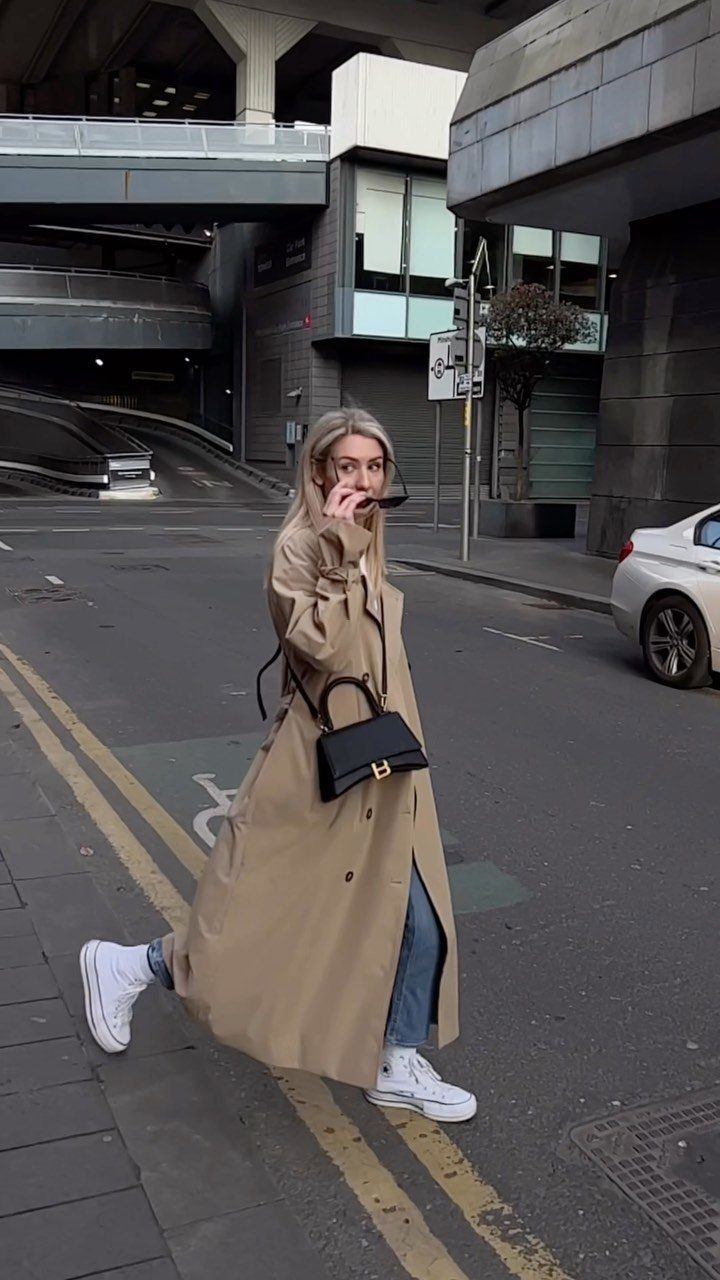 A woman wearing a long tan trench coat and blue jeans standing on a city street.
