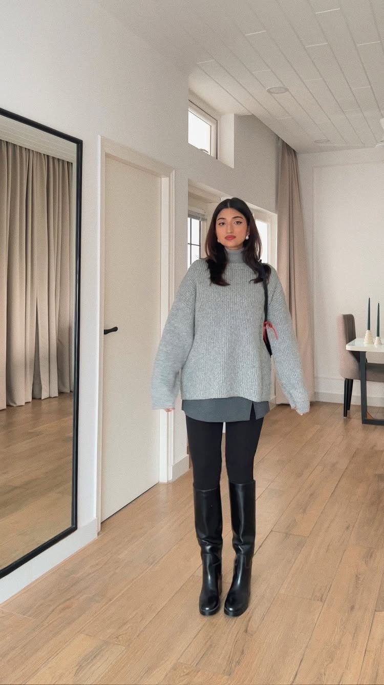 A woman wearing a grey turtleneck sweater, black leggings, and tall black boots.