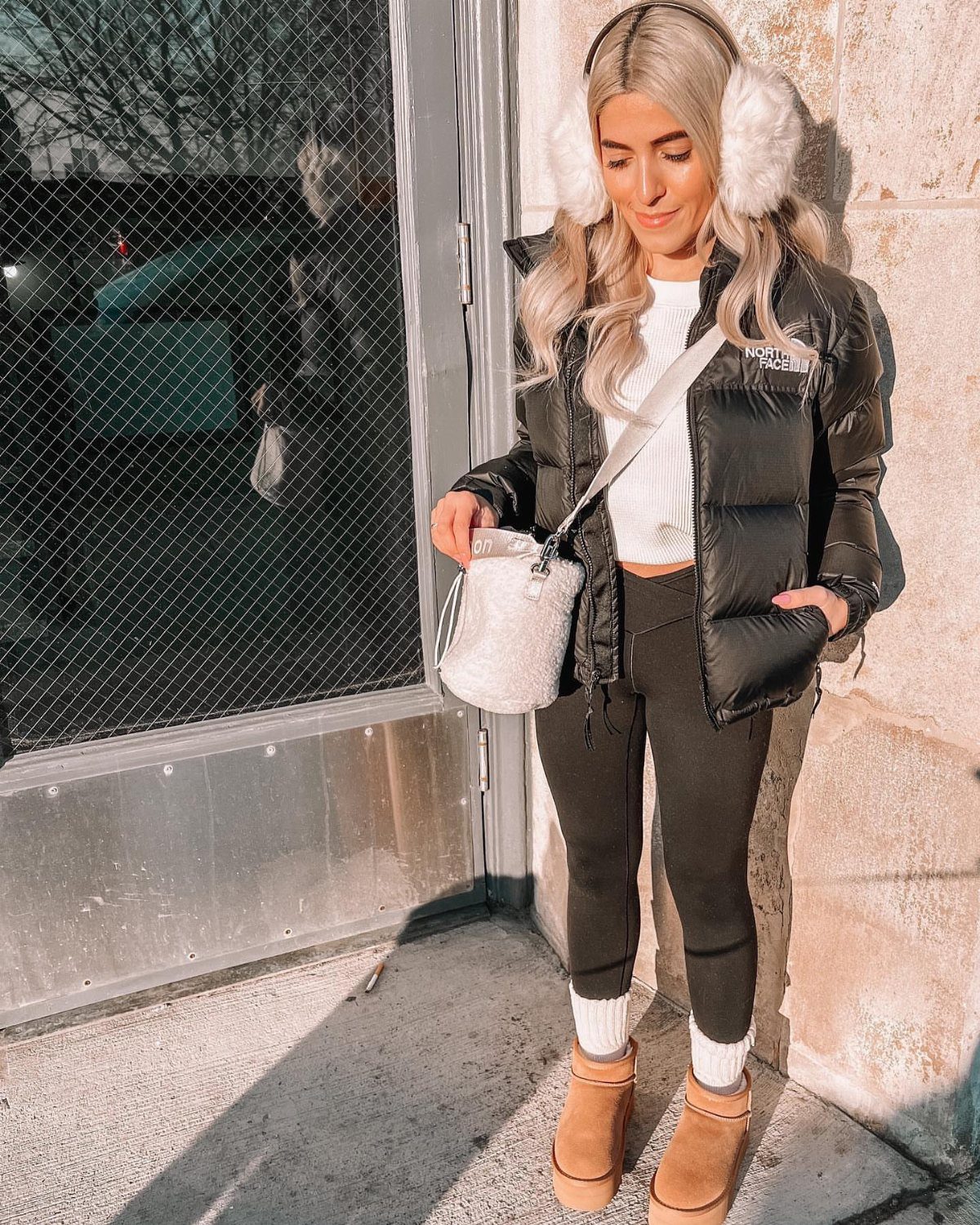 A woman in a black puffer jacket and black leggings standing outdoors by a stone wall.