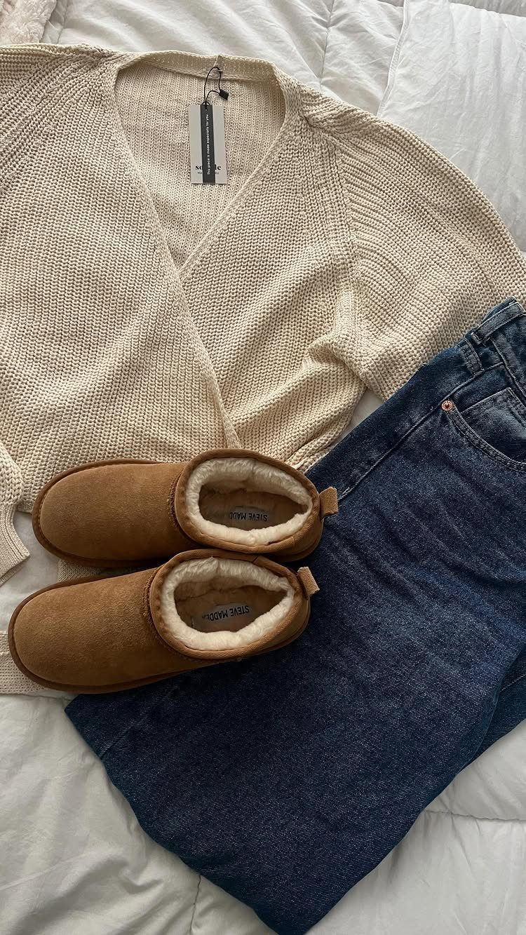 Cream knit cardigan, blue jeans, and brown suede slippers laid out on a white bedspread.