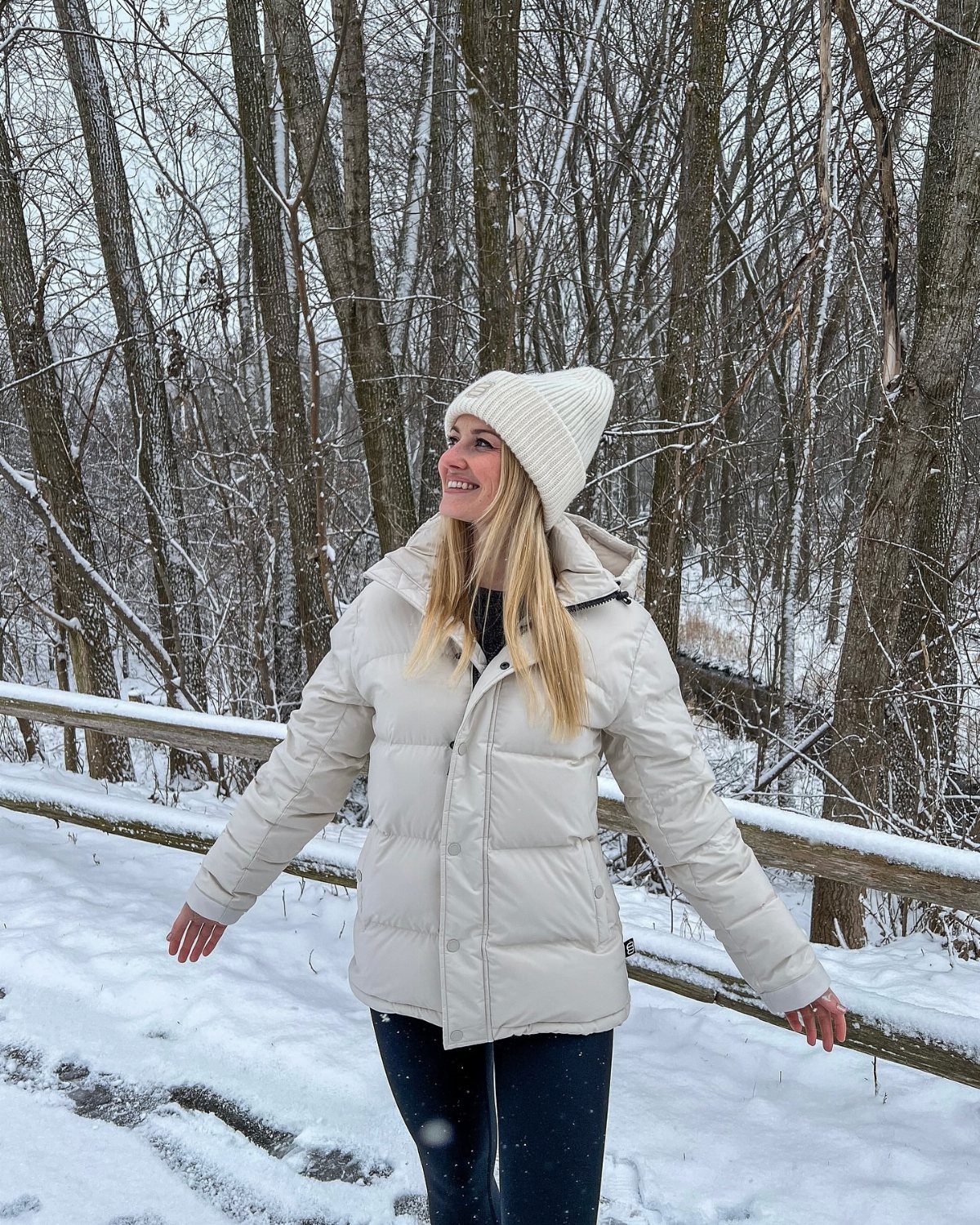 A smiling woman wearing a white beanie and cream puffer jacket in a snowy forest.