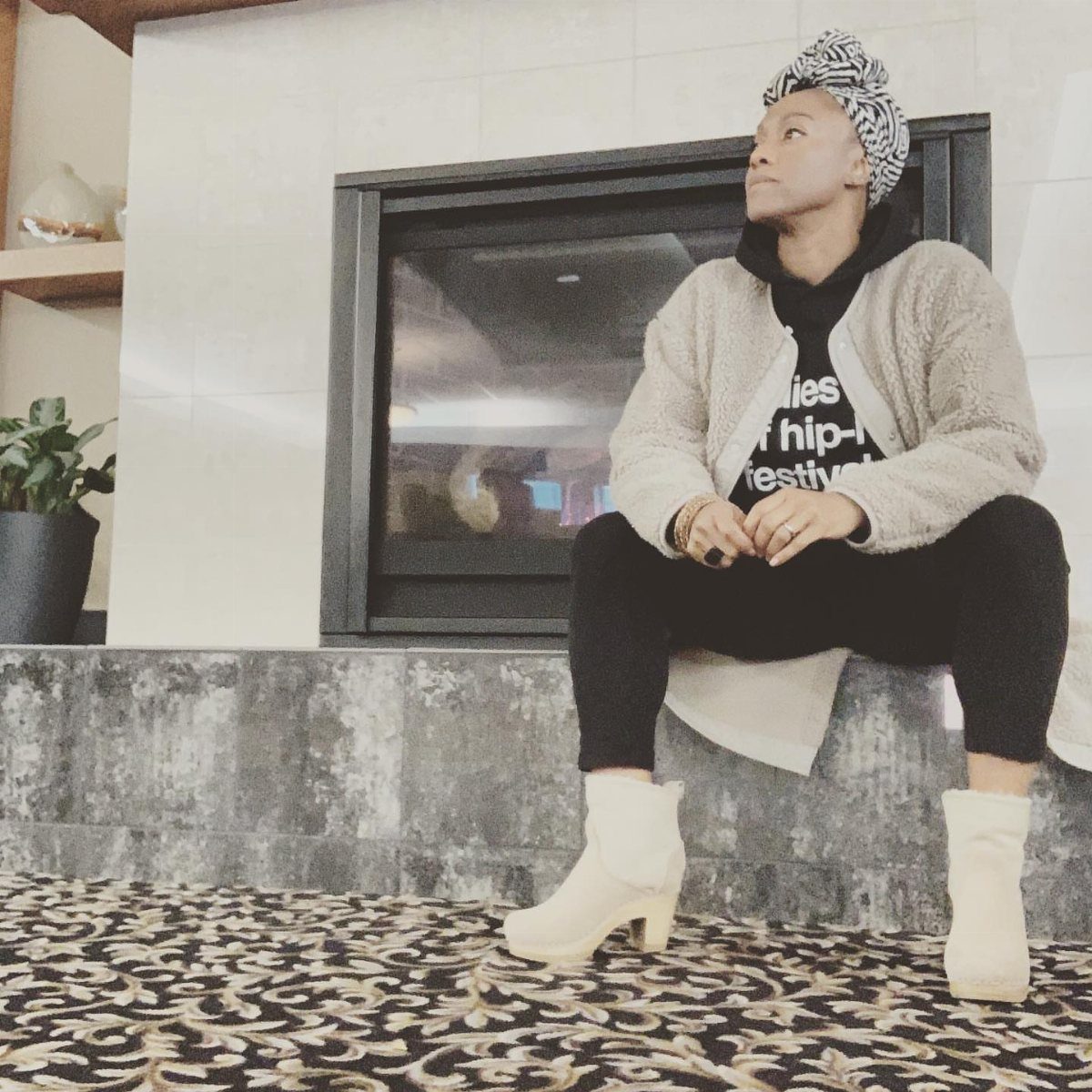 A person wearing a fuzzy jacket, black leggings, and white boots sitting by a fireplace.
