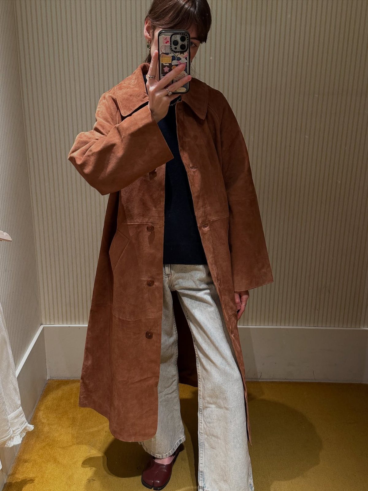 A person wearing a long brown suede coat and light denim jeans taking a mirror selfie.