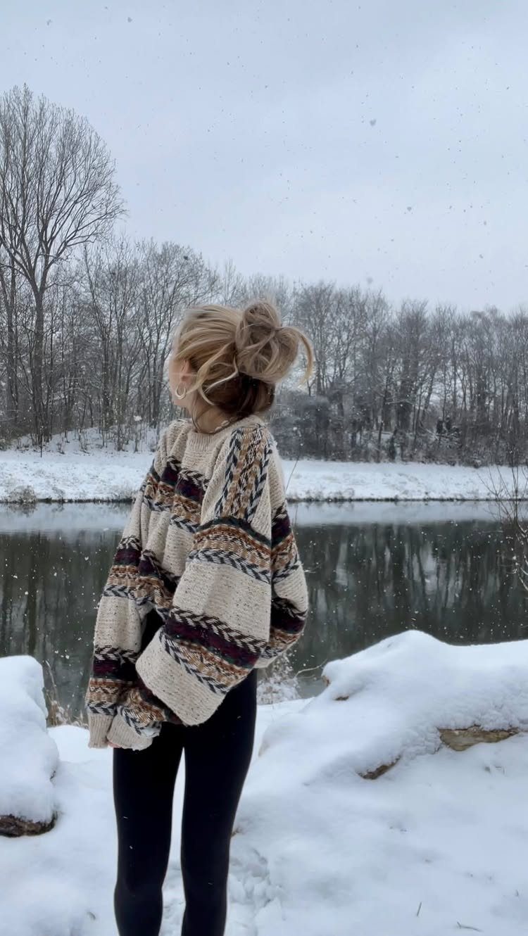 A woman in a patterned knit sweater and black leggings stands in a snowy landscape by a river.