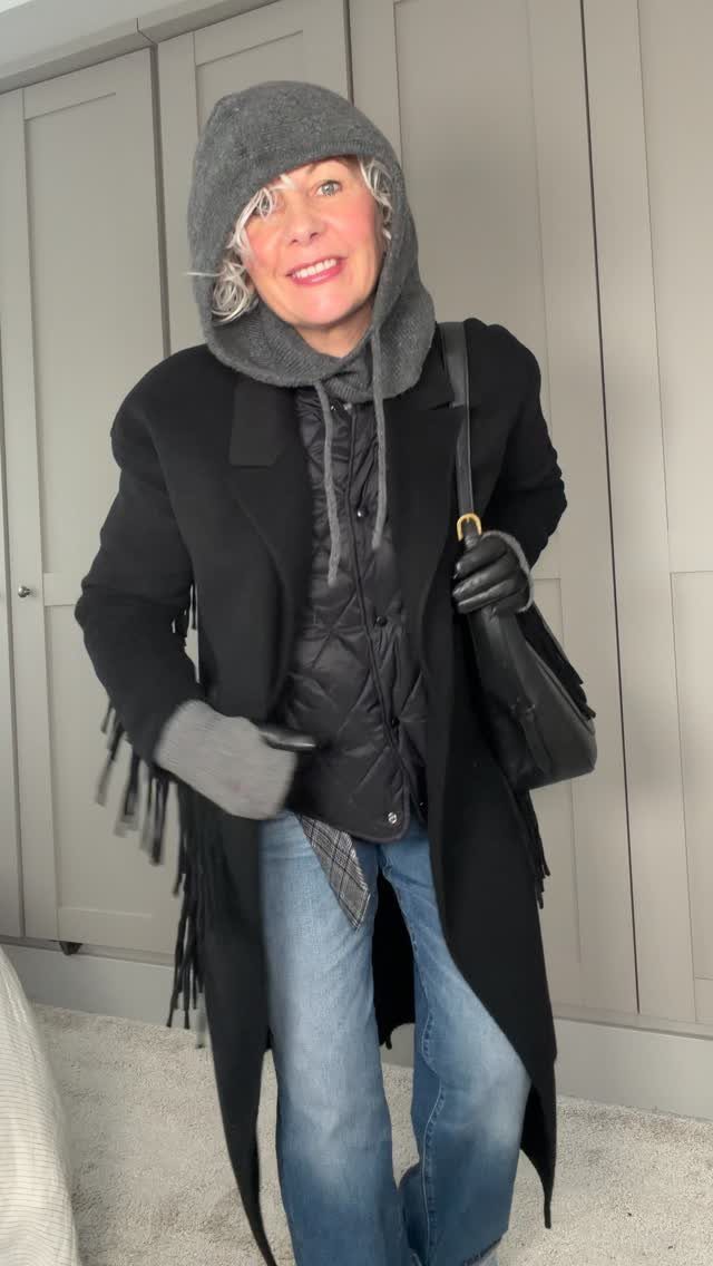 A woman wearing a black structured coat, quilted jacket, grey hood, and blue jeans.
