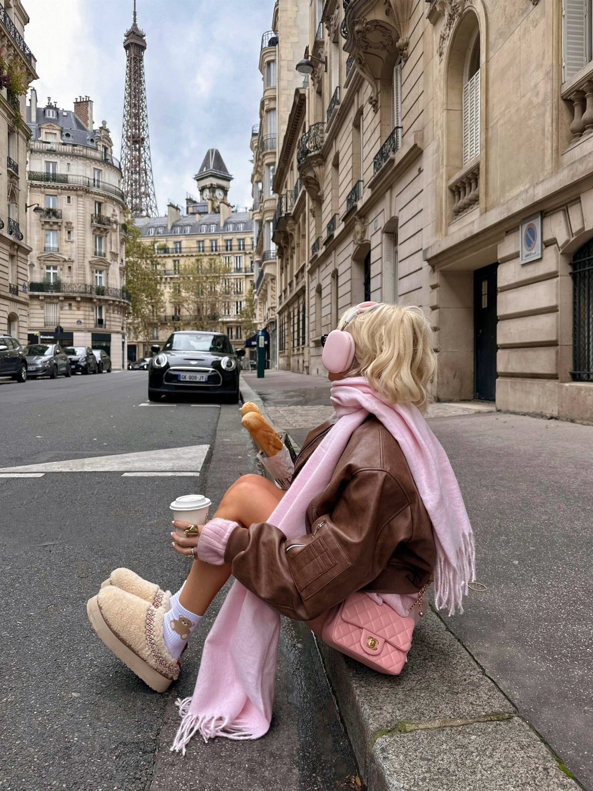 Blonde woman in a brown leather jacket and pink accessories sitting on a Parisian street curb.