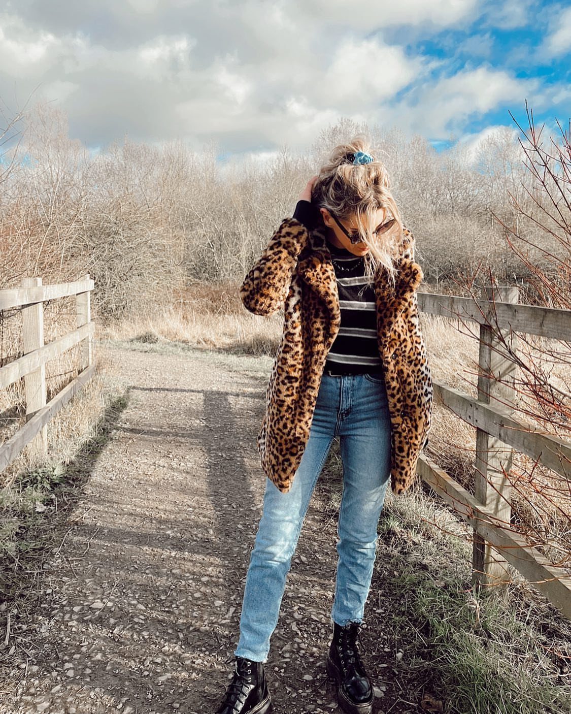 Woman wearing a leopard print coat, striped sweater, and blue jeans standing on a path.