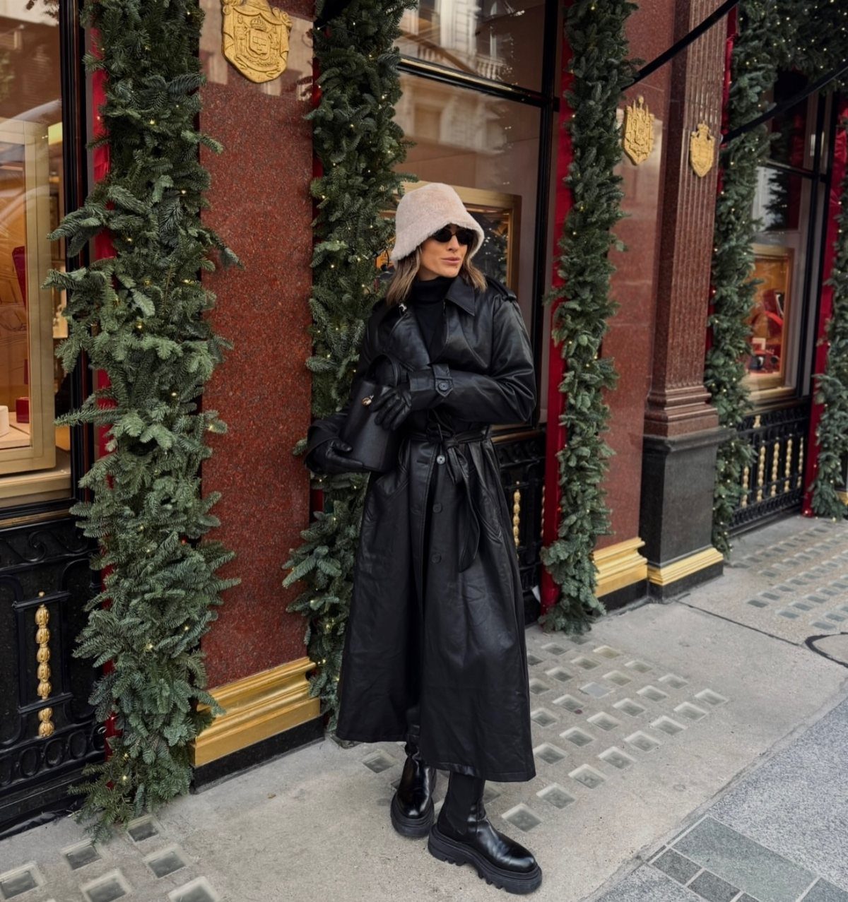 Woman in a long black leather coat, black turtleneck, and pink bucket hat standing outdoors.