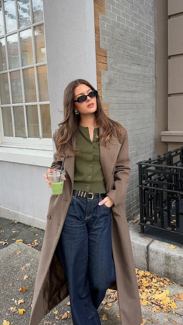 A woman wearing a long brown coat, olive shirt, and blue jeans standing outdoors.