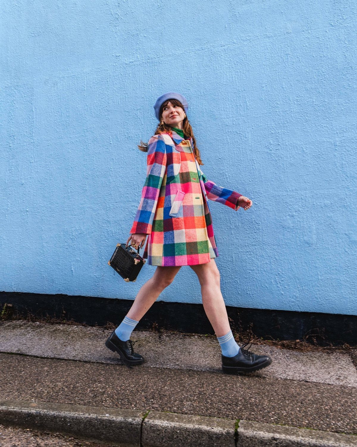 A woman walking in a vibrant multi-colored plaid coat against a solid blue wall.