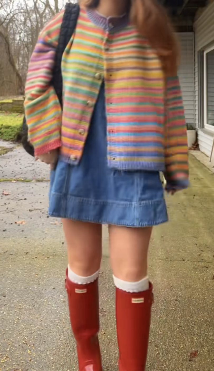Person wearing a colorful striped cardigan, denim skirt, and bright red tall rain boots.