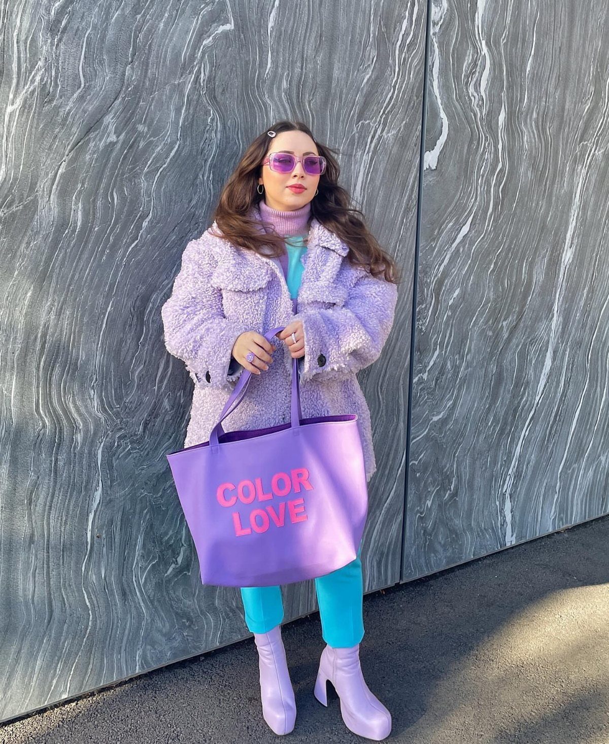 Woman wearing a lavender teddy coat, blue trousers, and pink boots holding a purple tote bag.