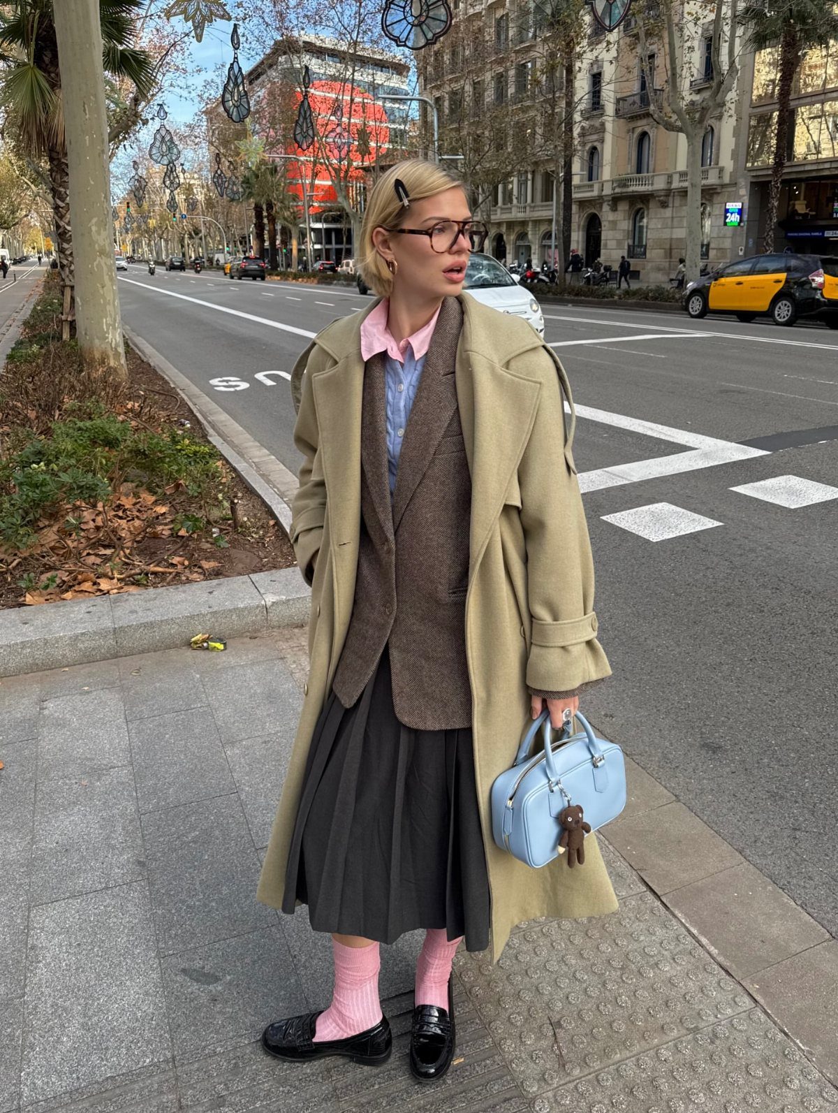 A blonde woman in a tan coat and pleated skirt standing on a city sidewalk.
