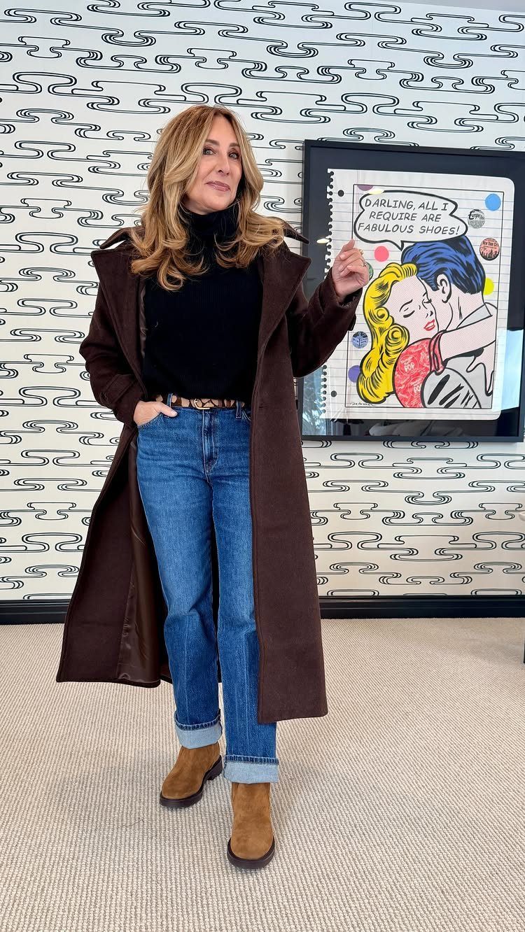 Woman in a long brown coat, blue jeans, and tan suede boots standing indoors.