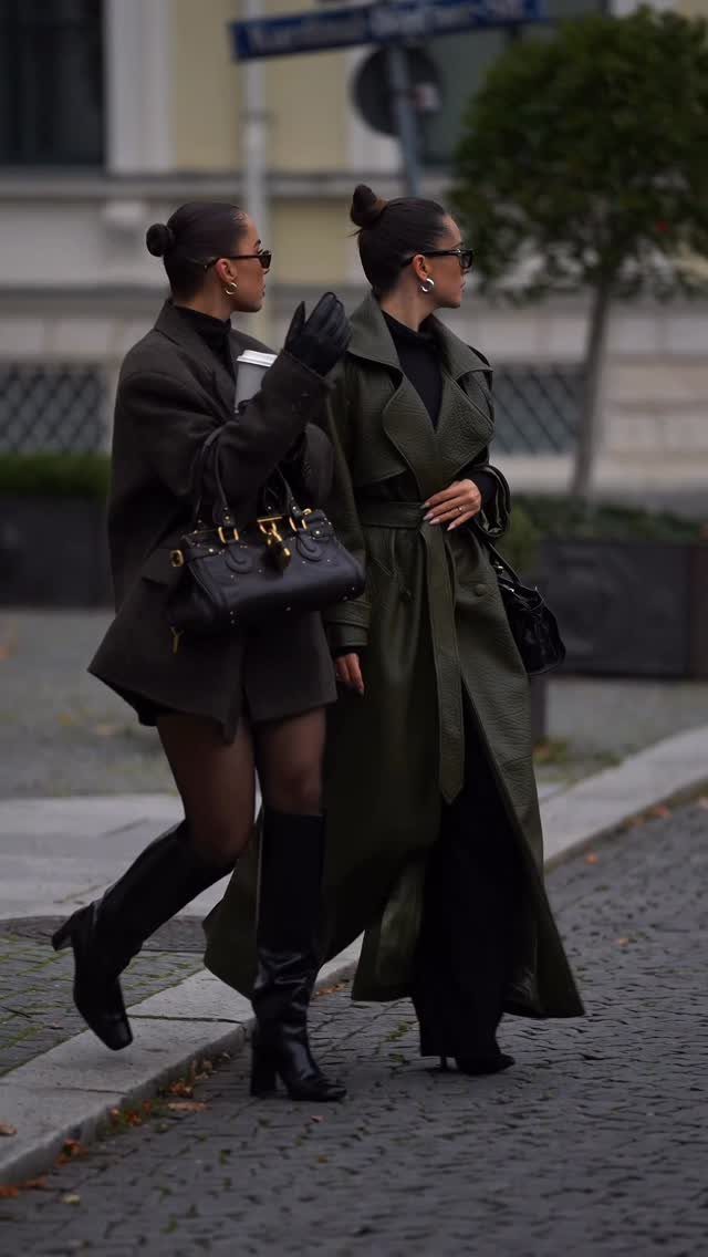 Two women walking down a street wearing long olive green coats and tall black boots.