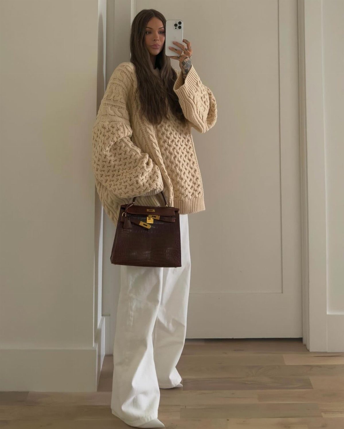 Woman in a beige textured oversized sweater and white wide-leg trousers holding a brown handbag.