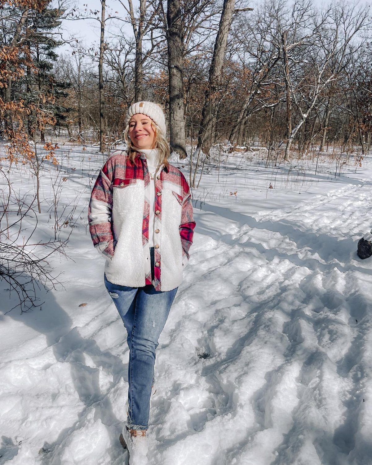 A woman in a red and white plaid sherpa shacket standing in a snowy forest.