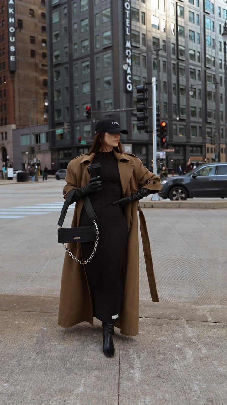 Woman in a tan trench coat and black dress standing on a city street.