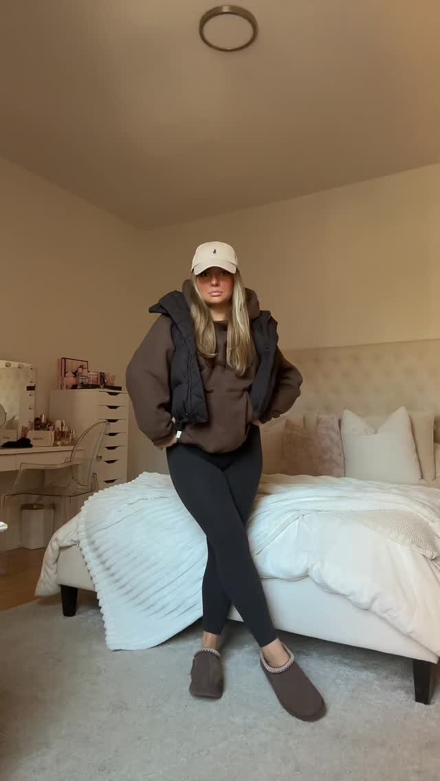 A woman posing in a bedroom wearing a brown hoodie, black puffer vest, and black leggings.