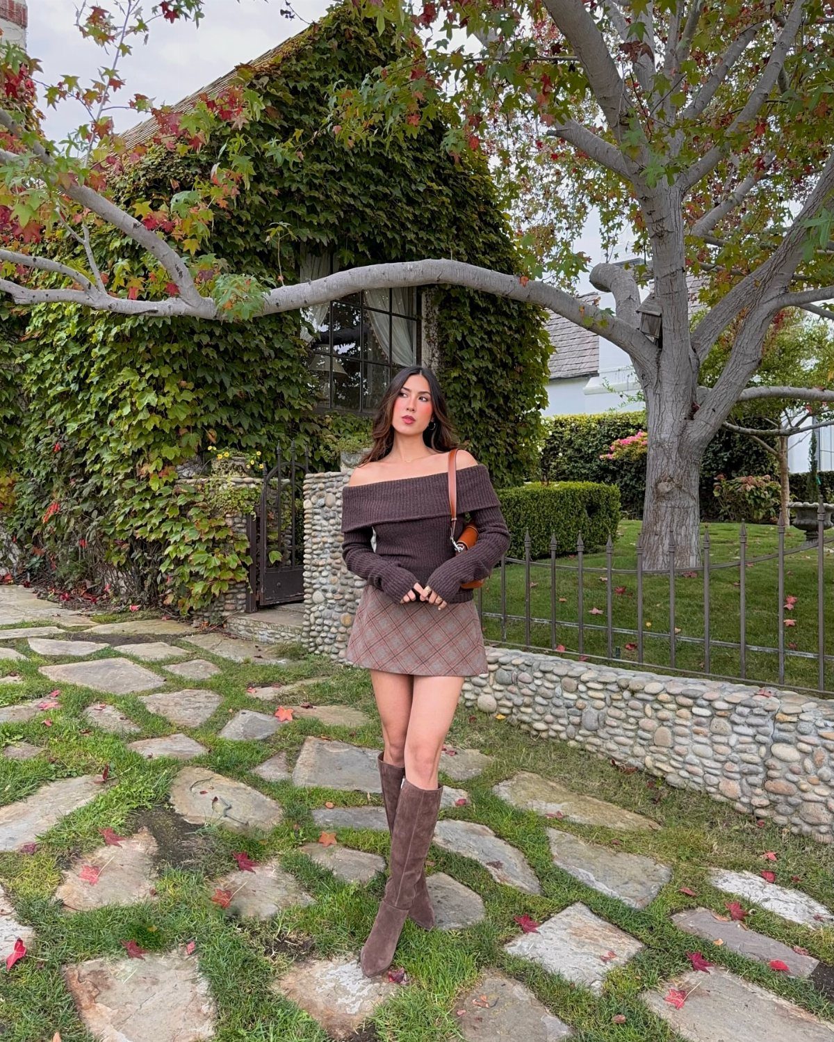 Woman wearing a brown off-the-shoulder sweater and a brown plaid mini skirt standing outdoors.