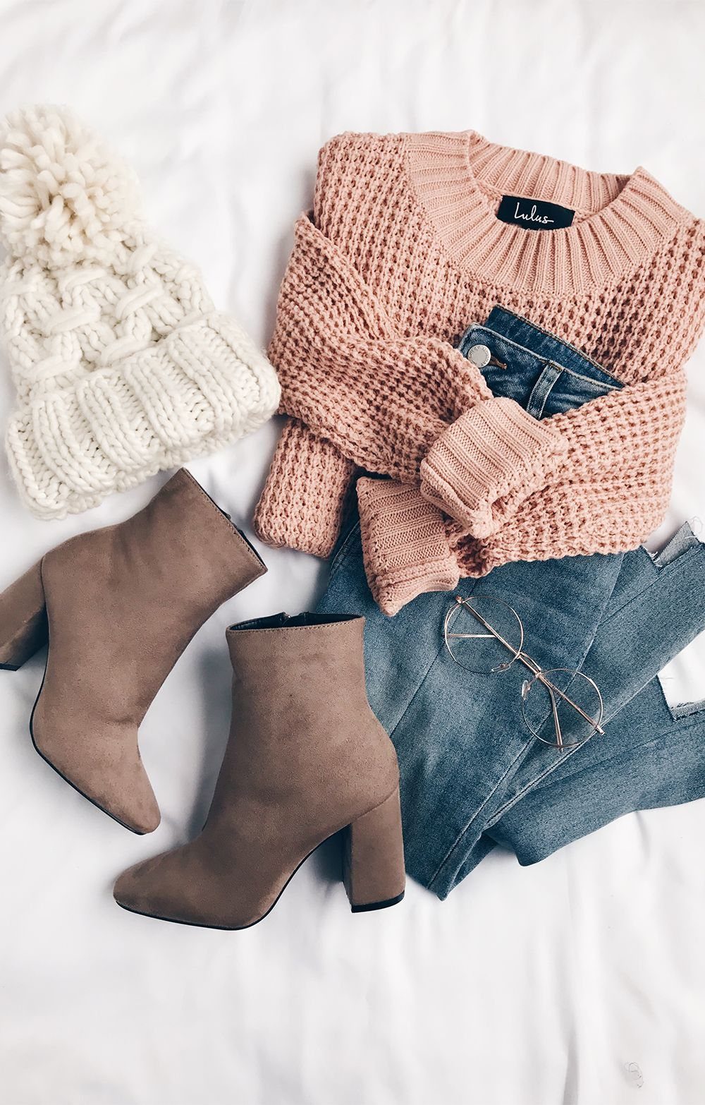 Brown suede ankle boots laid next to a pink sweater, blue jeans, and a white beanie.