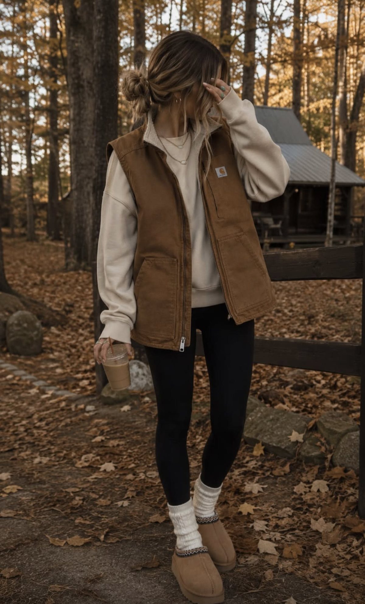 Woman wearing a tan fleece-lined vest, cream sweatshirt, and black leggings in an autumn forest.