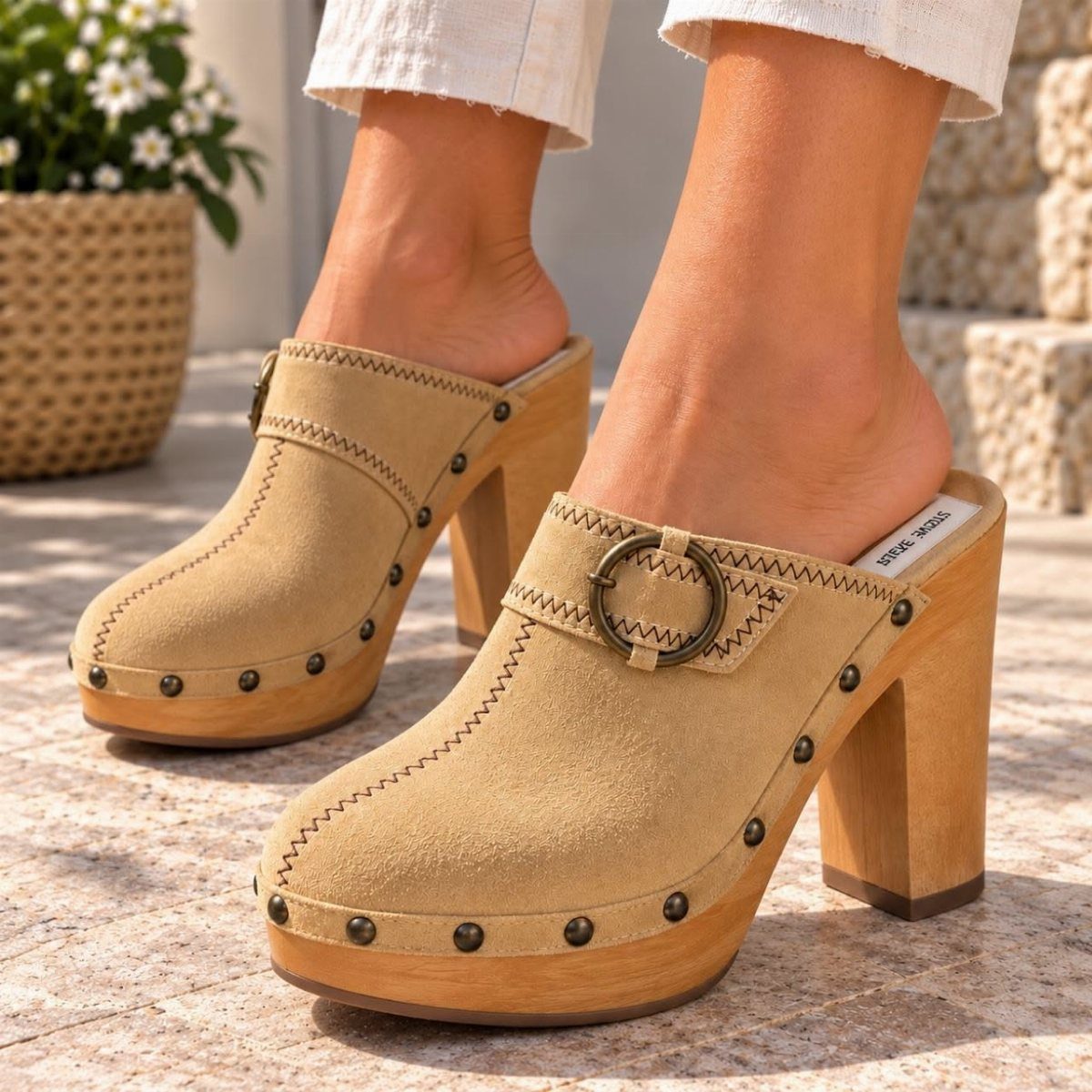 Tan suede platform clogs with chunky wooden heels and metal studs worn on stone ground.