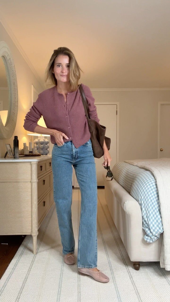 A woman posing in a bedroom wearing a mauve cardigan and light blue wide-leg jeans.
