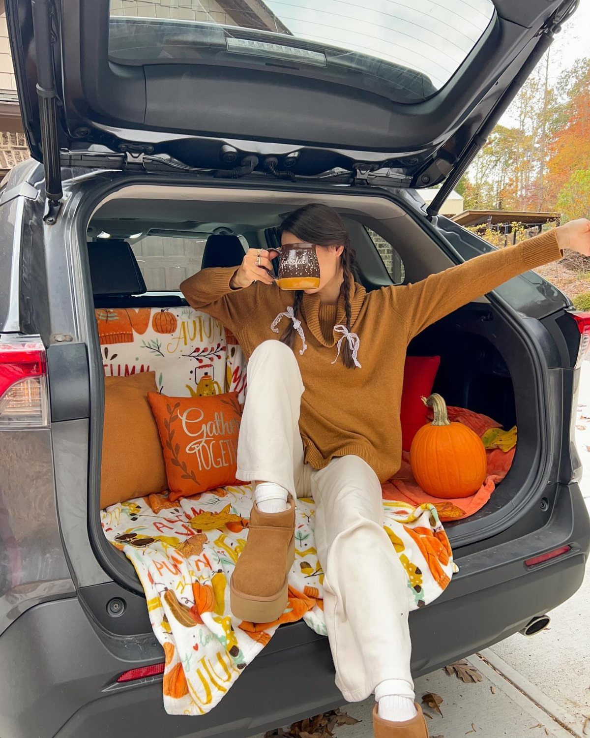 Woman in a brown sweater and white pants sitting in a car trunk with autumn decor.