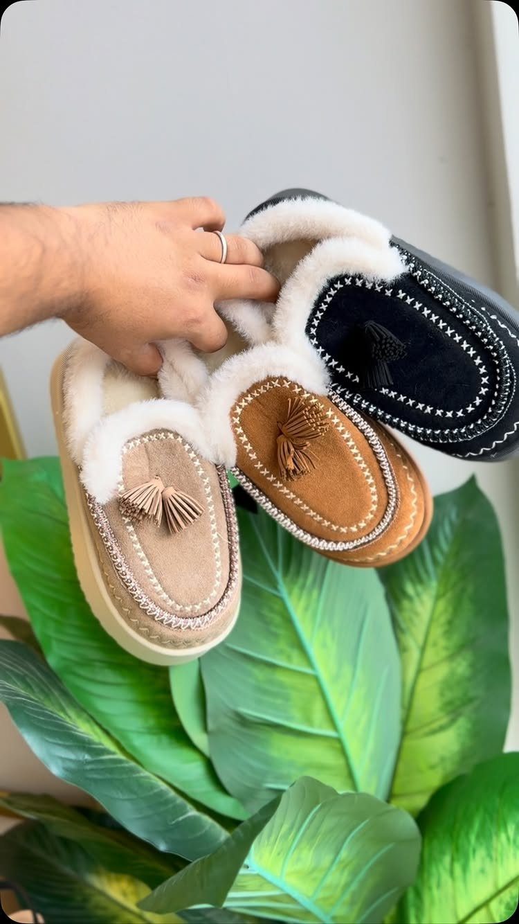 A hand holding three pairs of fuzzy tassel slides in beige, tan, and black colors.
