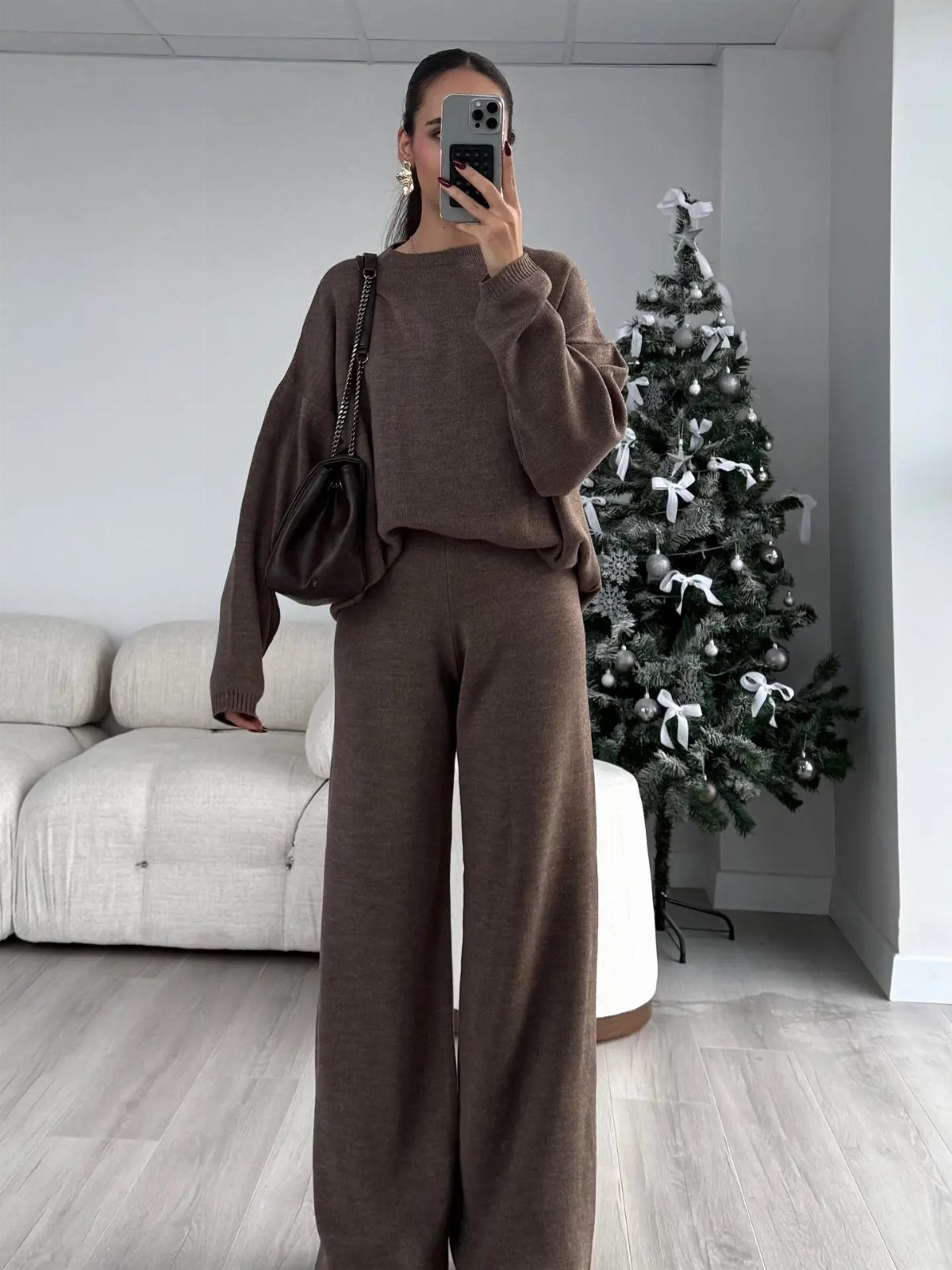 Woman in a brown knit pullover and wide-leg pants standing near a Christmas tree.