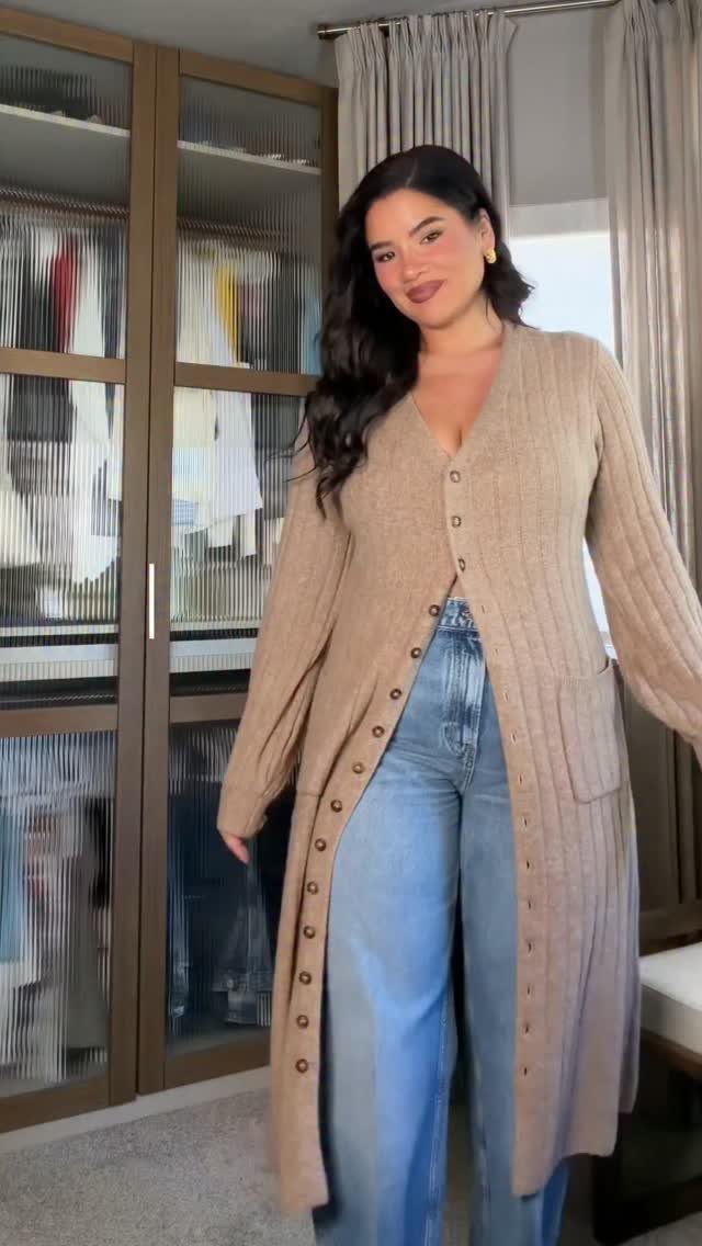 A smiling woman wearing a long tan ribbed knit cardigan and blue wide-leg jeans.