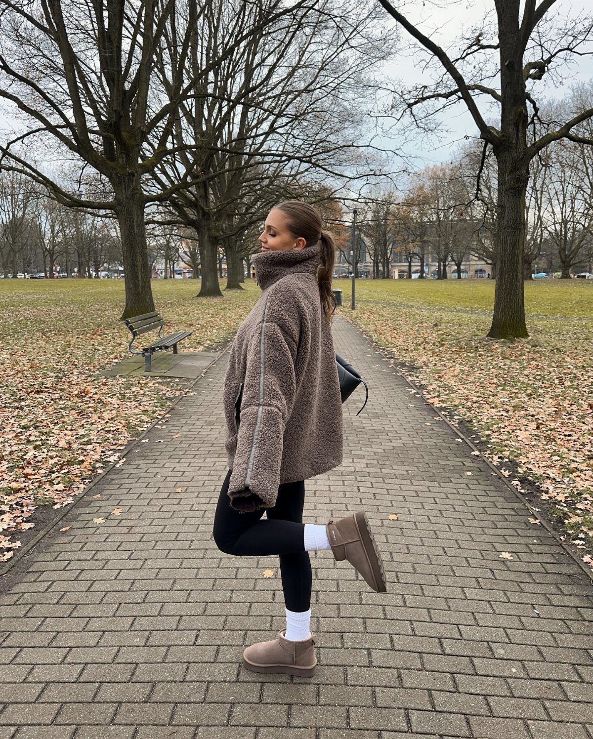 A woman wearing a brown teddy bear jacket and black leggings stands on a park path.