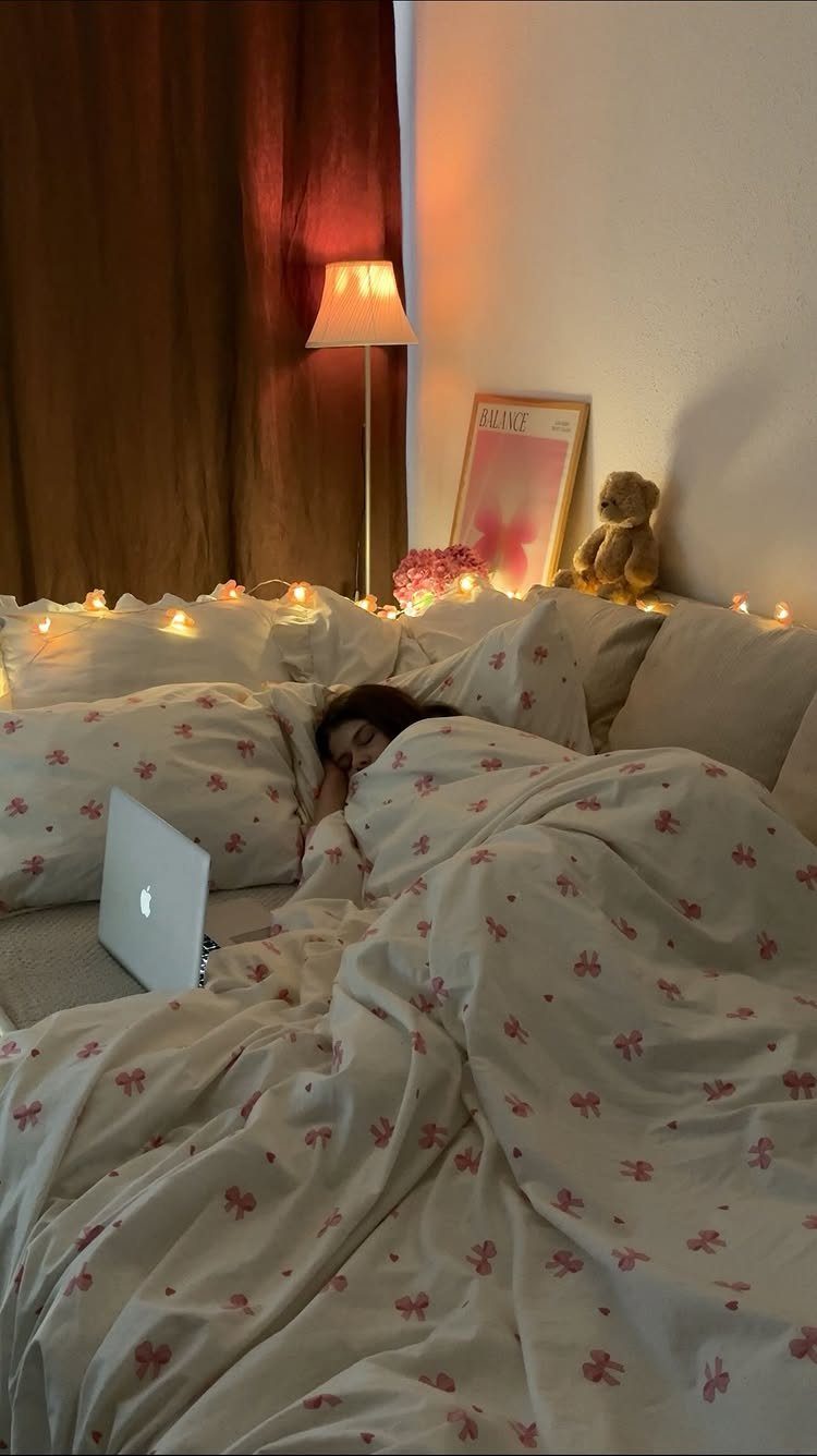 A person sleeping under floral blankets in a cozy, dimly lit bedroom with fairy lights.