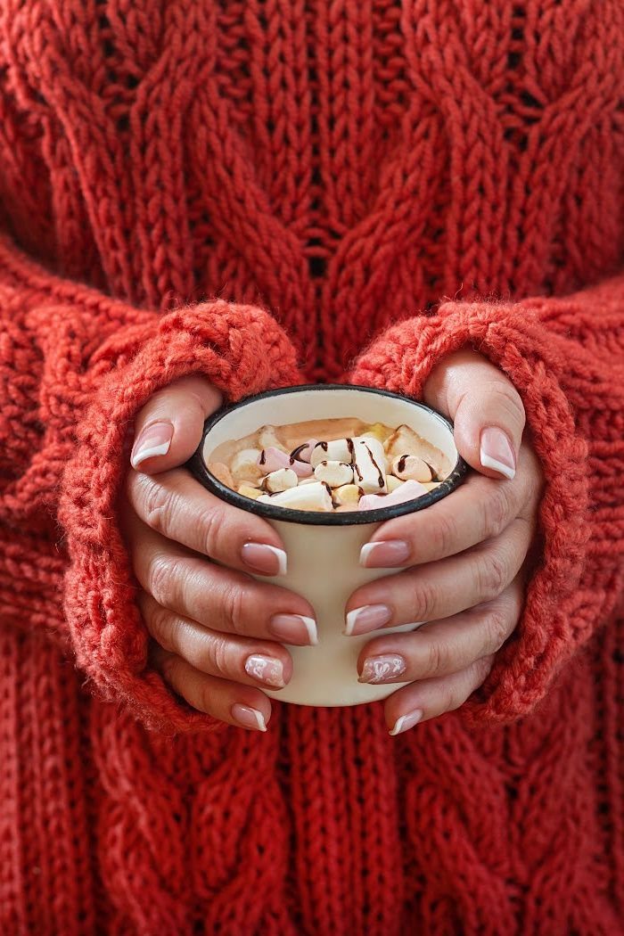 Hands in an orange knit sweater holding a mug of hot chocolate with marshmallows.