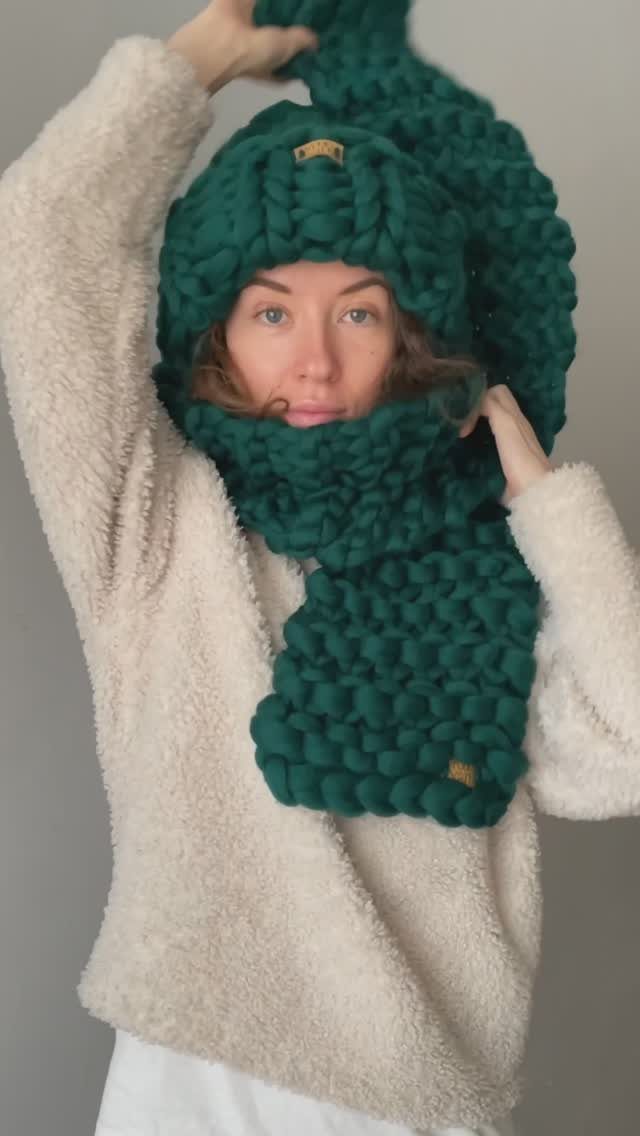 A woman wearing a cream fleece sweater and a large dark green chunky knit scarf and hat.
