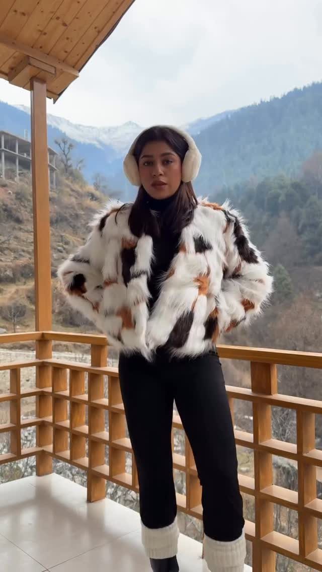 A woman wearing a patterned faux fur jacket and black clothing on a mountain balcony.