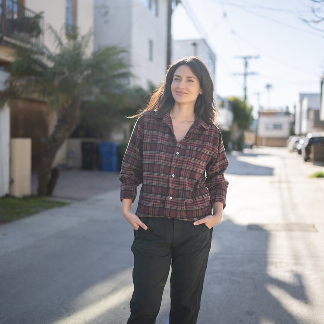 A woman wearing a red and black plaid flannel shirt standing on a sunny residential street.