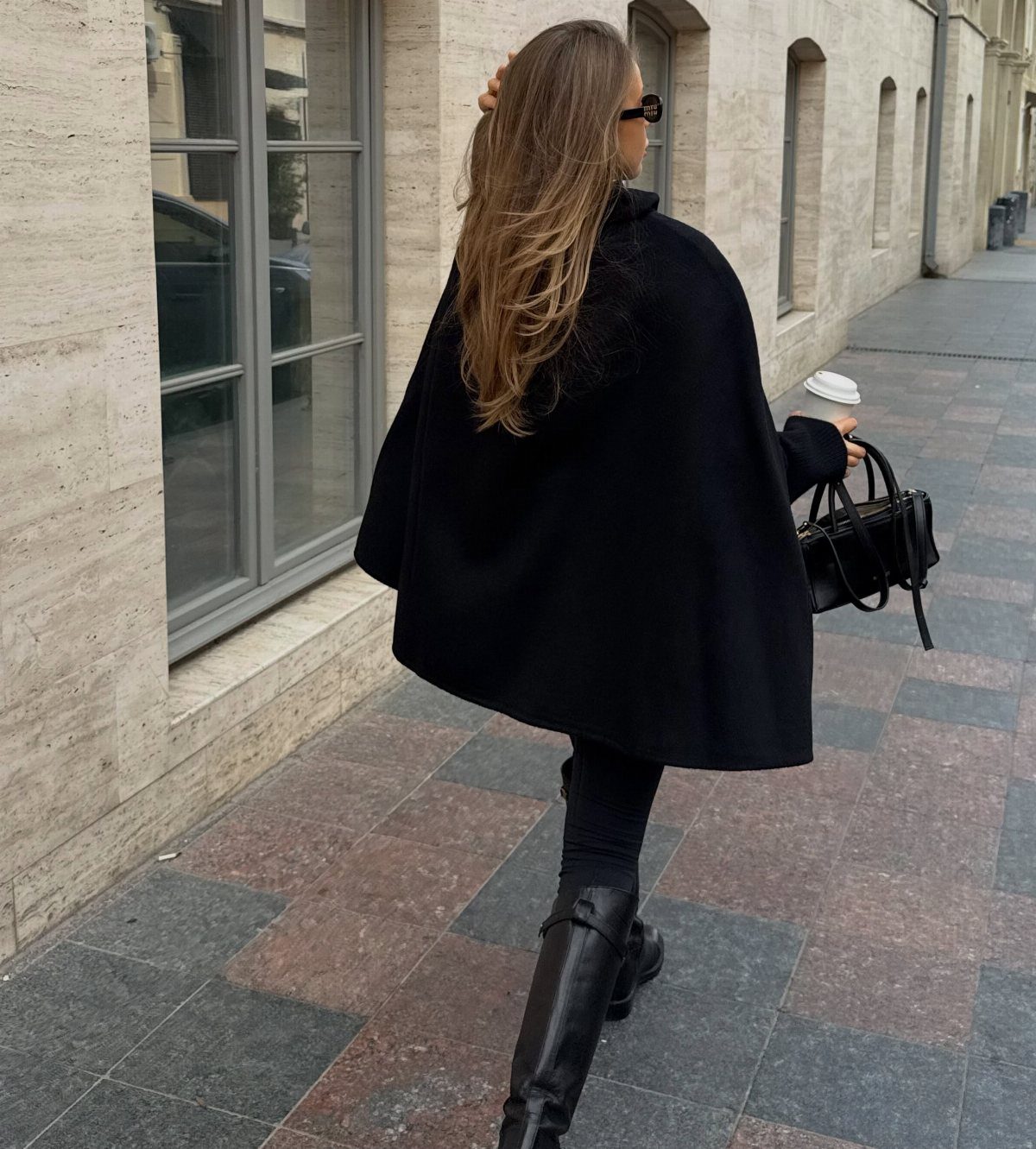 A woman walking on a city sidewalk wearing a black wool blend cape and tall boots.